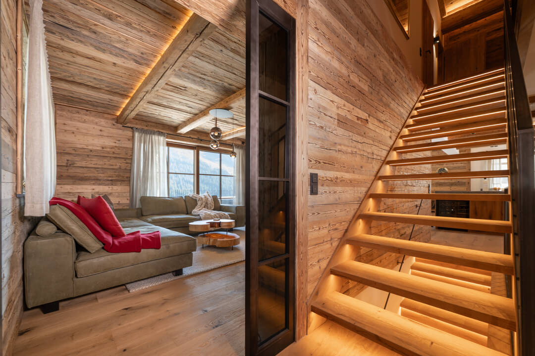 Alpine living room with dynamic ash burl coffee table, green leather sofa, red wool cushions, oak staircase with steel railing, oak flooring, and steel-look loft doors.