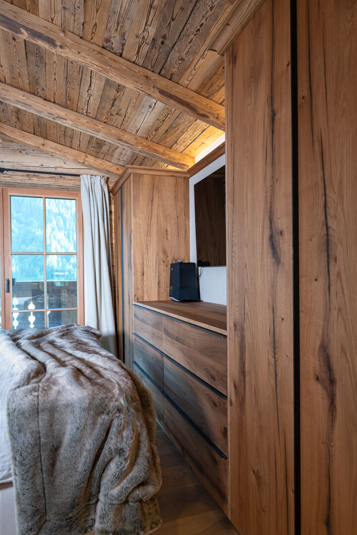 Alpine bedroom with built-in oak wardrobes, steel handles, cozy fur throw, light curtains, and reclaimed wood paneling.