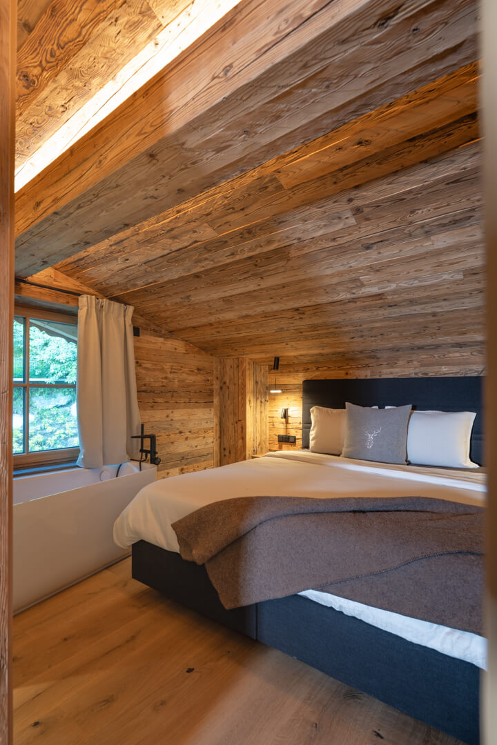 Cozy chalet bedroom with reclaimed wood ceiling and walls, corner bathtub, king-size bed, and Swarovski deer cushion.