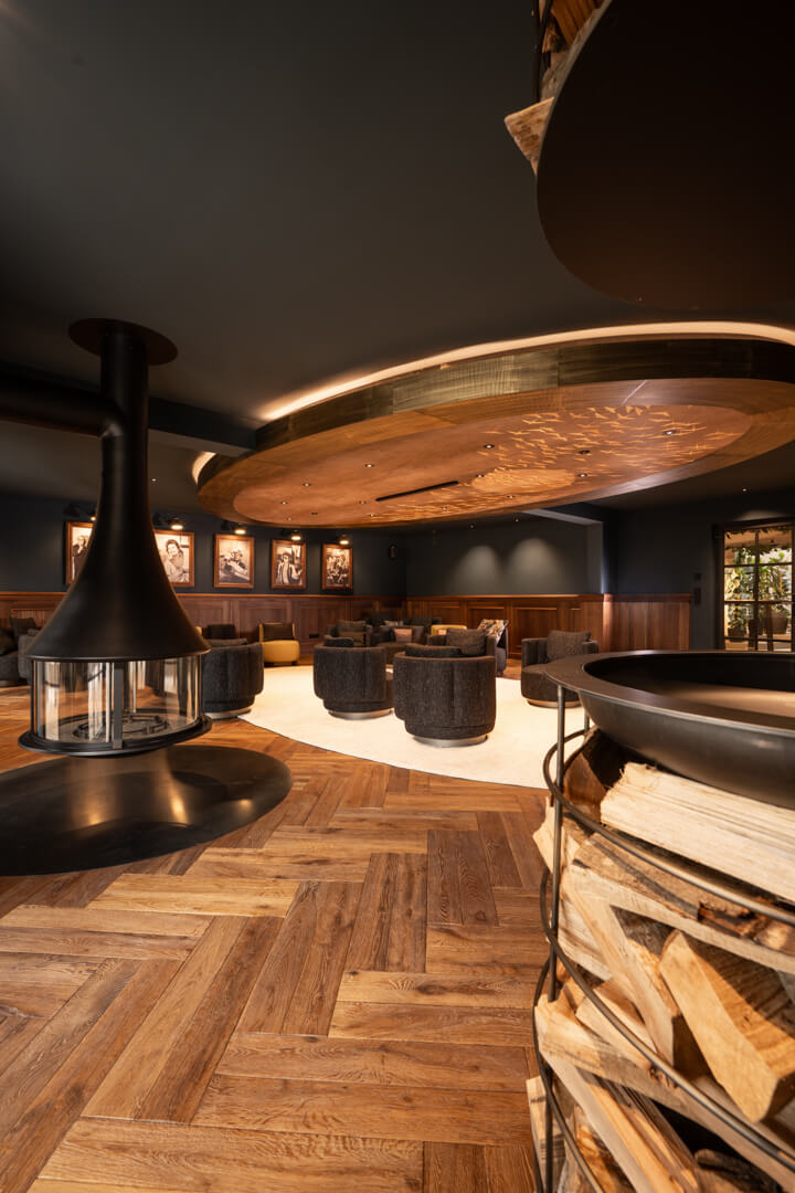 Fireplace lounge with dark interior, wood paneling, and herringbone flooring at Hotel Rössle. Light wooden carpet and champagne display centerpiece. Stacked wood and steel bowl.