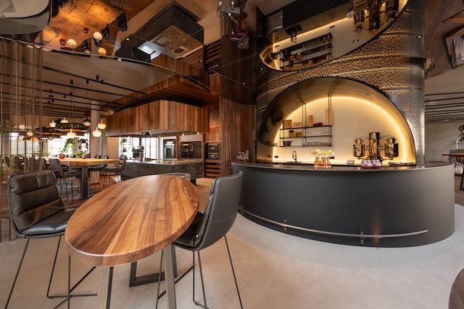 Warm walnut table, luxury grand kitchen with large stone island and wooden range hood, an imposing hotel bar with illuminated steel construction. 7,5 meter long oak table.