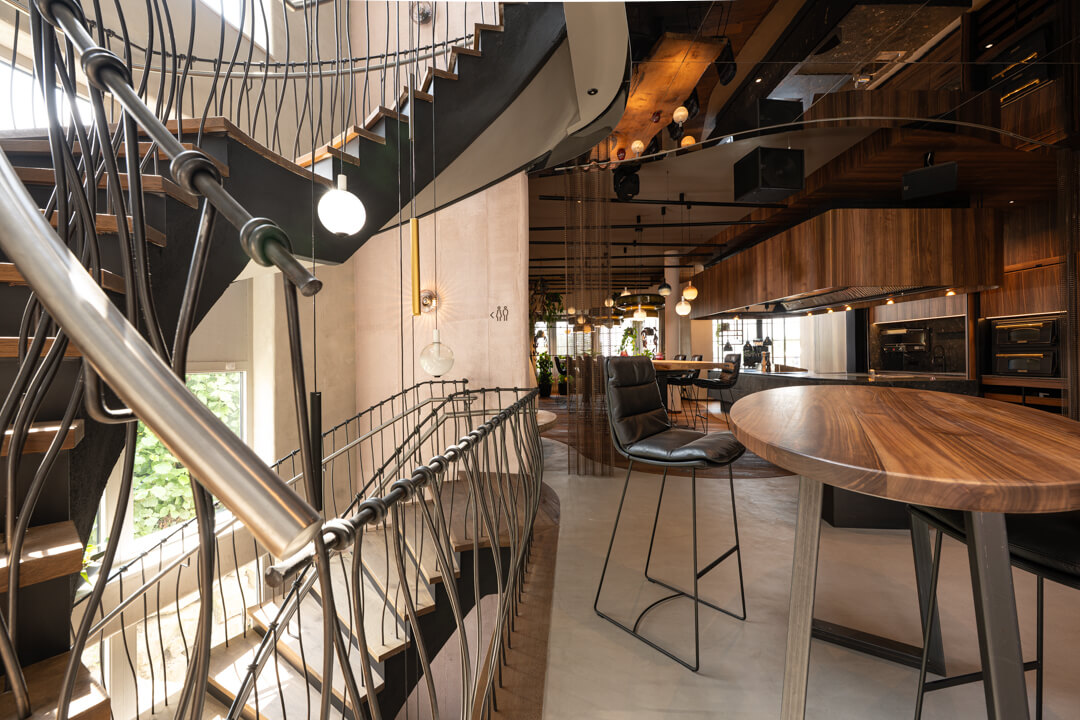 Oak staircase with dark wash, accompanied by a sculptural steel railing with organic lines and delicate knot details, alongside walnut bar tables with metal legs.